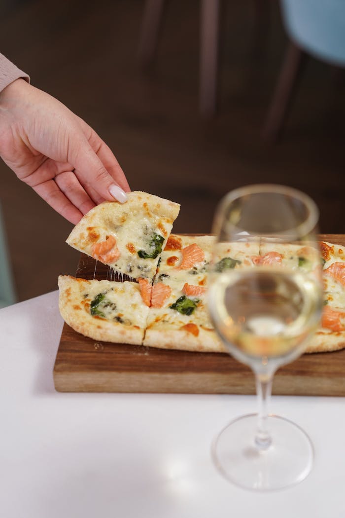 Hand picking a slice of gourmet Italian pizza with a glass of white wine on a wooden tray.